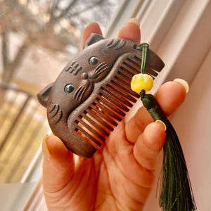 May include: A wooden comb shaped like a cat's face with green eyes and a yellow bead on a green tassel.