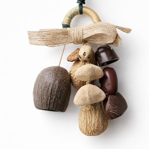 Peut inclure: Ornement décoratif suspendu fait à la main, composé de divers éléments naturels. Il comprend une grande pièce en forme de cloche texturée, brun foncé, ainsi que de plus petits éléments en forme de cloche et de graines, dans des tons bruns et beiges. Un nœud et une boucle en jute complètent le design.