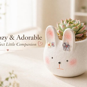 May include: A white ceramic bunny planter with a succulent plant. The planter features pink accents on the ears and cheeks, and small floral decorations. The succulent has green and pink leaves.