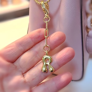 May include: A gold-colored charm with the letter "R" pendant, suspended from a chain. The charm is attached to a gold chain and clasp. The background is a light pink purse.