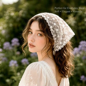 May include: A white lace headscarf with a floral pattern is worn by a person with long, wavy brown hair. The headscarf is triangular and covers the top of the head. The background is blurred with green foliage and purple flowers.