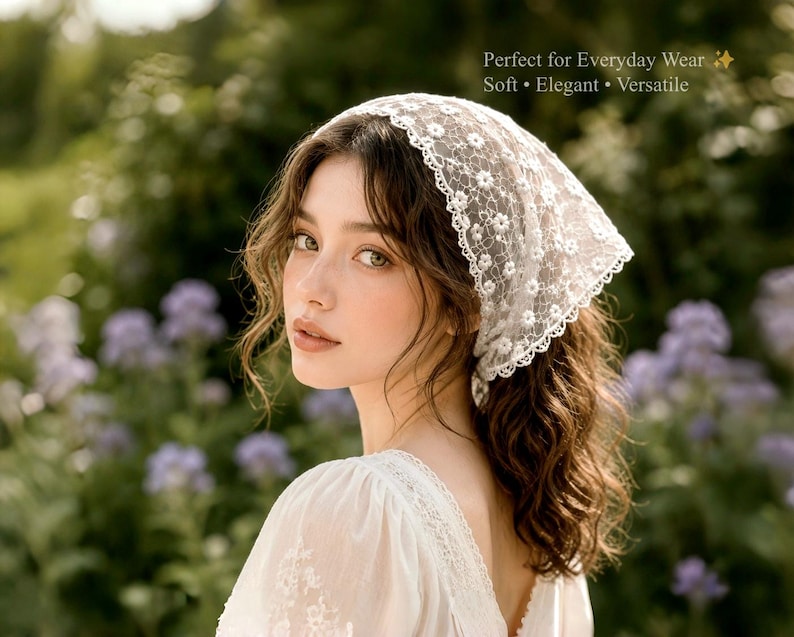 May include: A white lace headscarf with a floral pattern is worn by a person with long, wavy brown hair. The headscarf is triangular and covers the top of the head. The background is blurred with green foliage and purple flowers.