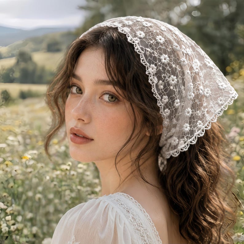 May include: A white lace headscarf with a floral pattern is worn over dark wavy hair. The headscarf is trimmed with a scalloped edge. The person is wearing a white dress and is in a field of flowers.