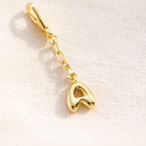 Op de afbeelding: Een goudkleurige bedel met de letter "A" in een afgerond, bubbelachtig lettertype. De bedel is verbonden met een ketting en een sluiting, geschikt om aan een armband of ketting te bevestigen. De bedel is gemaakt van glanzend, reflecterend metaal.