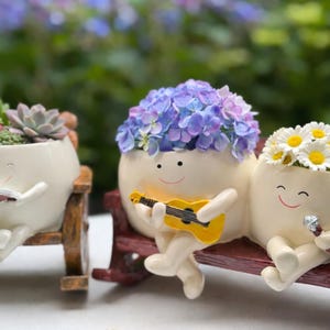 Puede incluir: Tres macetas de cerámica de color blanquecino con forma de personajes sonrientes. Una lee un libro, otra toca una guitarra amarilla y la tercera sostiene un micrófono. Cada maceta está llena de flores o suculentas, y están sentadas en bancos de madera.