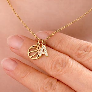 Peut inclure: Un collier doré avec un charm en forme de ballon de basket et un pendentif en forme de lettre "A". La lettre "A" est ornée de petits diamants transparents. Le collier est présenté sur une main.
