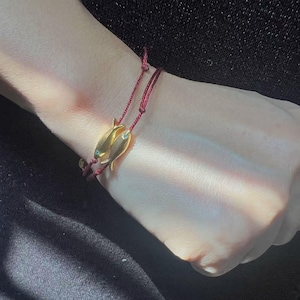 May include: A gold-colored bracelet featuring two fish-shaped charms. The charms are connected by a red cord. The bracelet is worn on a wrist, with the background out of focus.