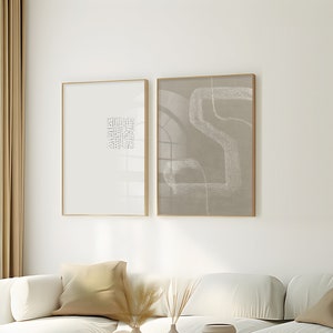 May include: Two framed abstract art prints in a minimalist living room setting. The prints are in gold frames and feature abstract line drawings in shades of grey. The prints are hanging on a white wall above a white sofa with beige cushions. A coffee table with a vase of dried pampas grass and other decorative objects is in front of the sofa.