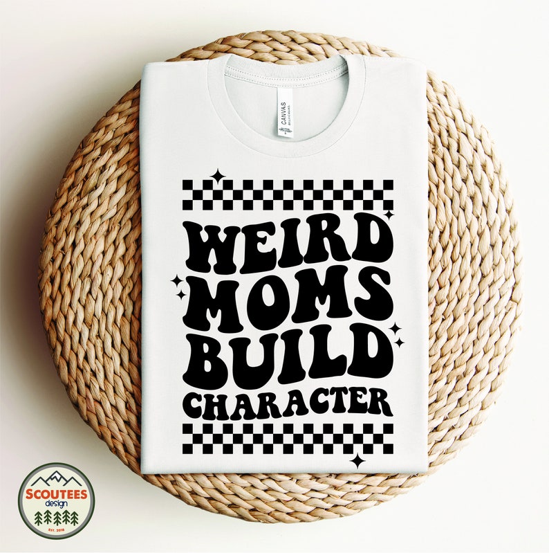 Op de afbeelding: Wit T-shirt met zwarte tekst die "Weird Moms Build Character" luidt met een geruit patroon boven en onder de tekst.
