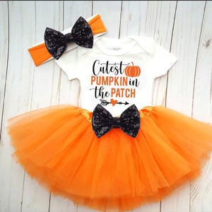 May include: A white baby bodysuit with the text "Cutest Pumpkin in the Patch" and an orange pumpkin graphic. The outfit includes an orange tutu skirt, a matching headband, and a black sequined bow.