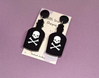 Peut inclure: Boucles d'oreilles noires en forme de bouteille avec un motif de tête de mort et d'os croisés blancs. Les boucles d'oreilles sont suspendues à des clous noirs. Le texte "Little Goat Designs" et "Handmade Jewellery" est visible.