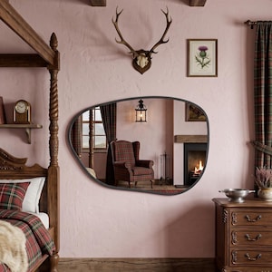 May include: An irregular, organic-shaped wall mirror with a thin, dark frame mounted on a light pink wall. The mirror reflects a room interior featuring a plaid armchair, a fireplace, and a hanging lantern, creating a clear, reflective surface.