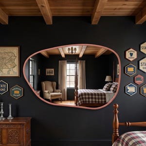 May include: An irregular, organic-shaped mirror with a muted pink frame mounted on a dark wall. The mirror reflects a bedroom interior featuring a wooden bed frame, a plaid comforter, and a beige armchair. The wall is decorated with hexagonal signs.