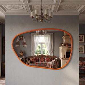 Può includere: Un grande specchio da parete dalla forma irregolare a ciottolo con una cornice arancione brillante. Lo specchio è montato su una parete grigia e riflette una stanza contenente un divano in legno con cuscini fantasia, un camino, un lampadario e tende bianche.