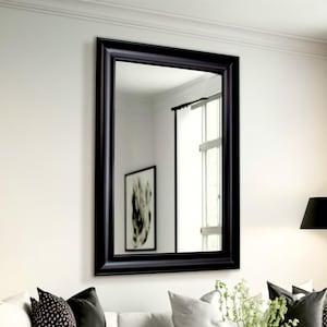 Pode incluir: Um espelho com moldura preta pendurado em uma parede branca acima de um sofá branco com almofadas de padrões preto e branco. Um abajur de chão preto com uma cúpula preta está no canto da sala.