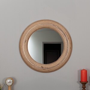 Può includere: Uno specchio rotondo con una cornice in legno naturale. La cornice ha un motivo a strati e a righe. Lo specchio riflette una stanza con una porta.