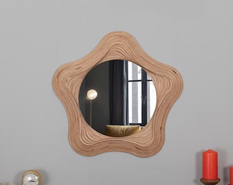 May include: A star-shaped wooden mirror with a wavy, layered frame. The mirror reflects a room with a window and a lamp.