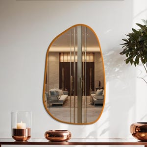 May include: An irregularly shaped mirror with a warm gold-toned frame hangs on a white wall. The mirror reflects a room with sofas and vertical glass panels. A copper-toned candle holder and bowl sit on a matching table.
