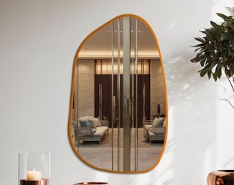 May include: An irregularly shaped mirror with a warm gold-toned frame hangs on a white wall. The mirror reflects a room with sofas and vertical glass panels. A copper-toned candle holder and bowl sit on a matching table.