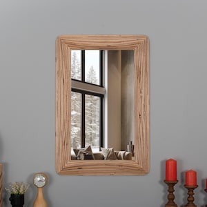 May include: A rectangular wooden framed mirror with a beveled edge. The mirror reflects a room with a large window and a view of trees. The mirror is hanging on a gray wall.