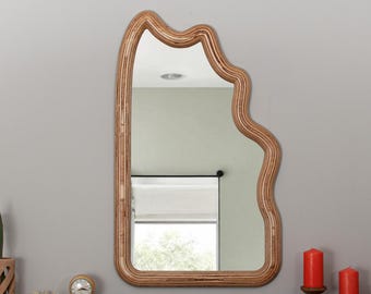 May include: A wooden framed mirror with a unique, wavy, organic shape. The mirror is hanging on a light gray wall.