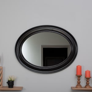 Puede incluir: Un espejo ovalado negro con un marco grueso cuelga de una pared gris claro sobre una repisa de madera con tres velas encendidas.