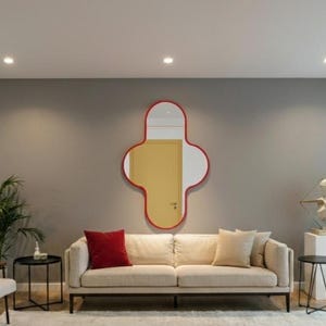 Könnte beinhalten: Ein modernes Wohnzimmer mit einem cremefarbenen Sofa, einem rot gerahmten Spiegel und einem weißen Teppich. Der Raum zeichnet sich durch eine neutrale Farbpalette mit grünen Pflanzen und einer Büstenskulptur aus und schafft so einen stilvollen und einladenden Raum.