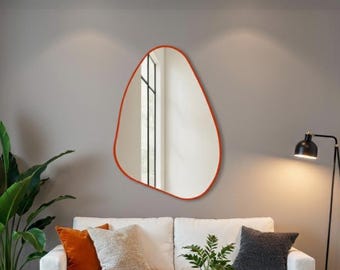 May include: A contemporary living room scene with a white sofa, cushions in orange and grey, and a wooden coffee table. A large, organic-shaped mirror with a red frame hangs on the wall. A black floor lamp and a potted plant are also visible.