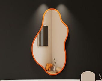 May include: An orange-edged, freeform mirror is mounted above a white and wood-effect dressing table. The mirror reflects a room with a window and fruit. A small black stool sits below the table, and a small mirror and diffuser are on the table.