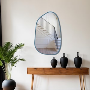 Blue framed asymmetrical wall mirror in a mid-century modern hallway above wooden console with black ceramic vases and indoor plant.