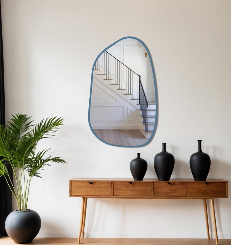 Blue framed asymmetrical wall mirror in a mid-century modern hallway above wooden console with black ceramic vases and indoor plant.
