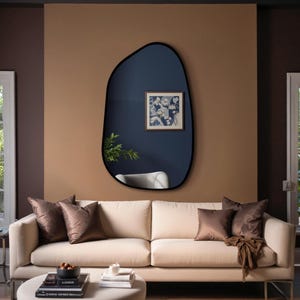 Black framed irregular wall mirror above beige sofa in a cozy modern living room with dark accent wall and natural lighting