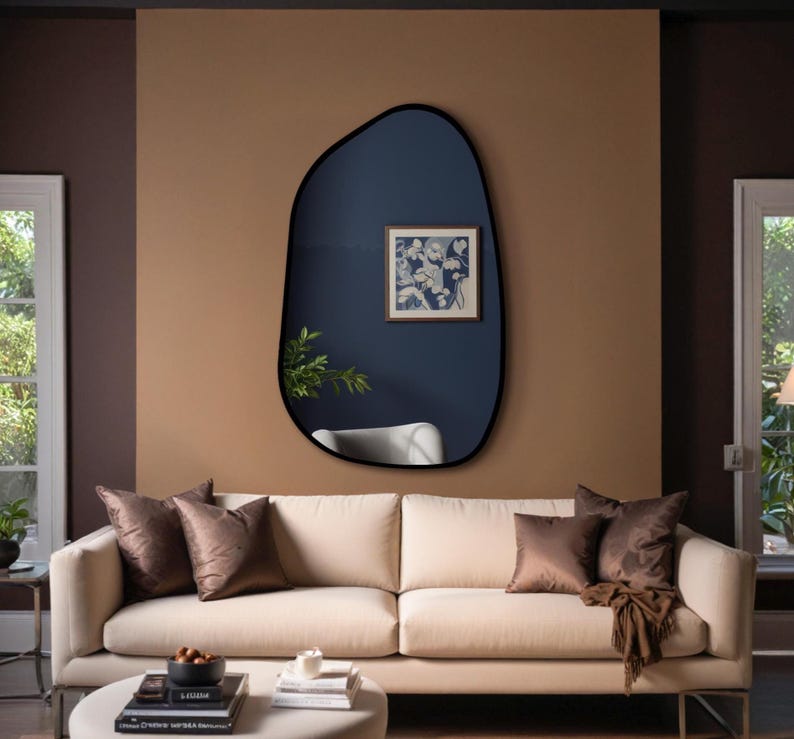Black framed irregular wall mirror above beige sofa in a cozy modern living room with dark accent wall and natural lighting