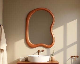 Puede incluir: Escena de baño moderno con un gran espejo de color cobre de forma orgánica. Debajo del espejo hay un lavabo blanco con grifos de cobre. Una toalla cuelga a la izquierda y una pequeña planta está a la derecha.