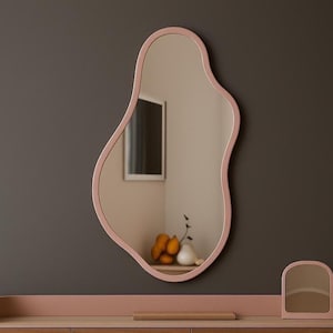 May include: A large, organically shaped mirror with a soft pink frame hangs on a dark gray wall. The mirror reflects a room with a window and a small arrangement of fruit and vases. A matching pink vanity with a small mirror sits below.