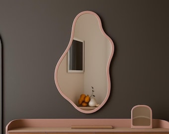 May include: A large, organically shaped mirror with a soft pink frame hangs on a dark gray wall. The mirror reflects a room with a window and a small arrangement of fruit and vases. A matching pink vanity with a small mirror sits below.