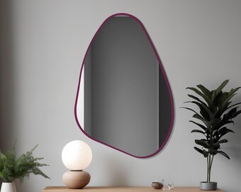 May include: An irregularly shaped mirror with a deep magenta frame hangs on a gray wall. A modern, minimalist design, the mirror reflects a room. A white globe lamp and a potted plant sit below the mirror on a wooden console.