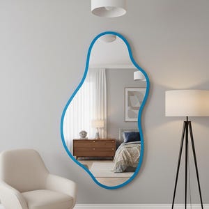 May include: An abstract-shaped mirror with a bright blue border is mounted on a grey wall. The mirror reflects a bedroom scene with a bed, chest of drawers, and lamp. A white armchair is in the foreground.