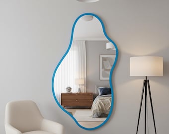 May include: A large, irregularly shaped mirror with a vibrant blue frame hangs on a gray wall. The mirror reflects a bedroom scene with a bed, dresser, and lamp. A white armchair sits in the foreground.