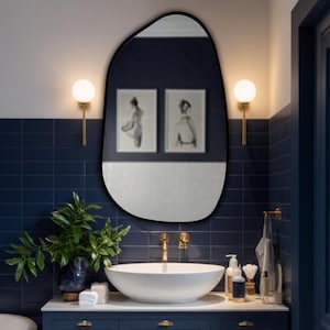 Black framed asymmetrical mirror above white vessel sink in a navy blue bathroom with gold fixtures and elegant wall lighting