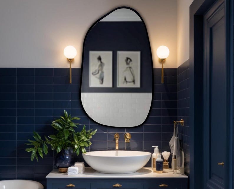 Black framed asymmetrical mirror above white vessel sink in a navy blue bathroom with gold fixtures and elegant wall lighting