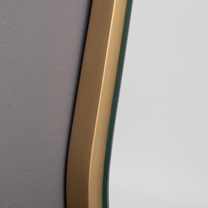 May include: A wall mirror featuring a smooth, matte gold-toned frame with a curved edge. The reflective glass surface is set within the frame, showing a clear reflection of a neutral-colored wall. The frame has a consistent metallic finish.