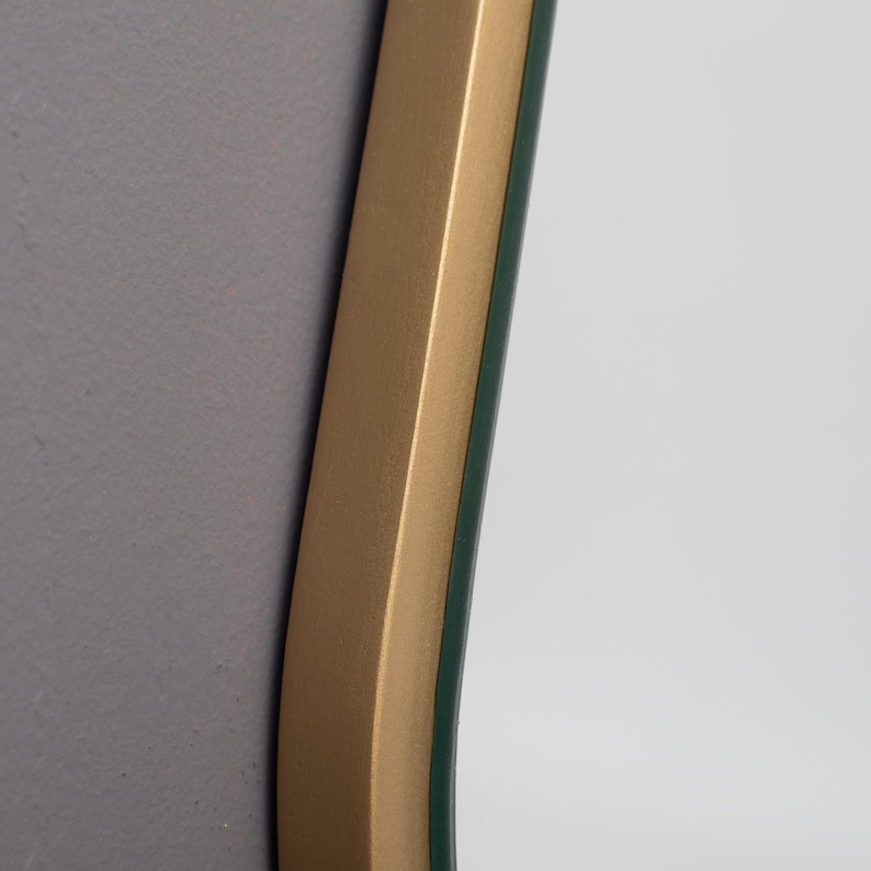 May include: A wall mirror featuring a smooth, matte gold-toned frame with a curved edge. The reflective glass surface is set within the frame, showing a clear reflection of a neutral-colored wall. The frame has a consistent metallic finish.