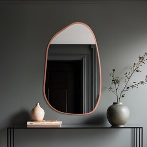 Puede incluir: Un espejo de pared de forma irregular con marco color cobre y sección superior blanca. El espejo está sobre una mesa negra con un jarrón, libros y una planta decorativa. La pared está pintada de color gris oscuro.