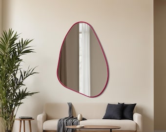 May include: A modern, irregularly shaped mirror with a vibrant magenta frame hangs on a neutral wall. The mirror reflects a window with white curtains. The room features a beige sofa, dark pillows, and a wooden coffee table, creating a minimalist aesthetic.