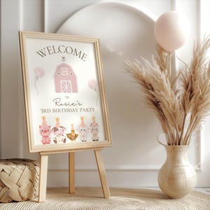 May include: A framed sign on an easel with the words "WELCOME" and "Rosie's 3RD BIRTHDAY PARTY". The sign features a pink barn illustration and cartoon farm animals. A vase of pampas grass and a pink balloon are also in the image.