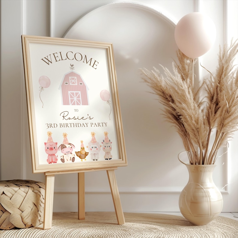 May include: A framed sign on an easel with the words "WELCOME" and "Rosie's 3RD BIRTHDAY PARTY". The sign features a pink barn illustration and cartoon farm animals. A vase of pampas grass and a pink balloon are also in the image.