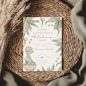 May include: A baby shower invitation with a green dinosaur hatching from an egg. The invitation has a watercolor background with green leaves and the text "RAWR! A LITTLE BOY IS Hatching SOON". The invitation also includes the date, time, and address for the shower.
