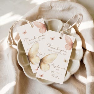 Peut inclure: Deux étiquettes cadeaux rectangulaires avec des illustrations de papillons et le texte "Thank you for coming to Sophia's 1st Birthday Party". Les étiquettes ont un fond crème avec des papillons aquarelles pêche et jaunes. Chaque étiquette est attachée avec de la ficelle naturelle.
