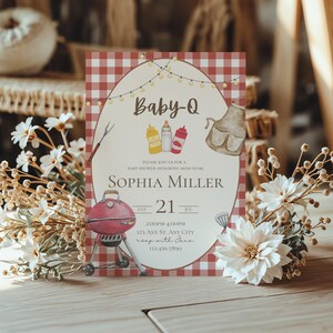Op de afbeelding: Baby-Q babyshower uitnodiging met een rood-witte geruite achtergrond. De uitnodiging bevat een barbecue-illustratie, tekst en decoratieve elementen. Witte bloemen en gedroogde bloemaccenten omringen de uitnodiging.